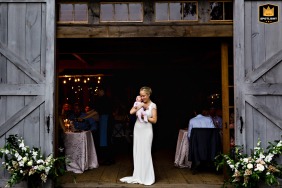 A heartwarming moment outside the intimate wedding venue, Primo Restaurant in Rockland Maine, showing the bride holding their newborn daughter in the opening of large barn doors on their special day.